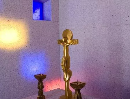 Eine goldene Skulptur steht im Raum, umgeben von zwei kleinen, dunklen Figuren. Bunte Lichtstrahlen fallen auf die Wände und schaffen eine ruhige Atmosphäre.