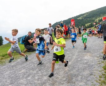 A group of children enthusiastically runs along a dirt path. In the background, spectators and a hilly landscape can be seen.