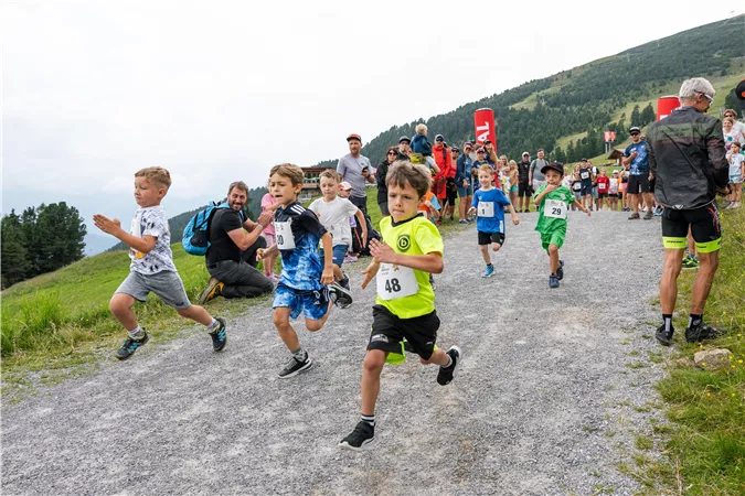 Eine Gruppe von Kindern läuft enthusiastisch einen Feldweg entlang. Im Hintergrund sind Zuschauer und eine hügelige Landschaft zu sehen.
