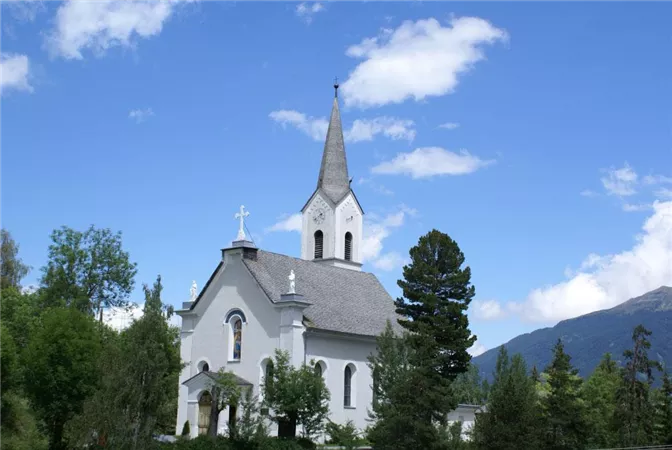 A white church with a sharp steeple against a blue sky. Surrounded by green trees and mountains.