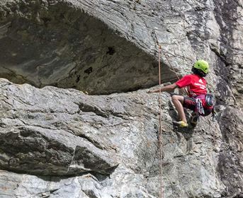 A climber in a red shirt ascends a steep rock. He is wearing a helmet and climbing gear.