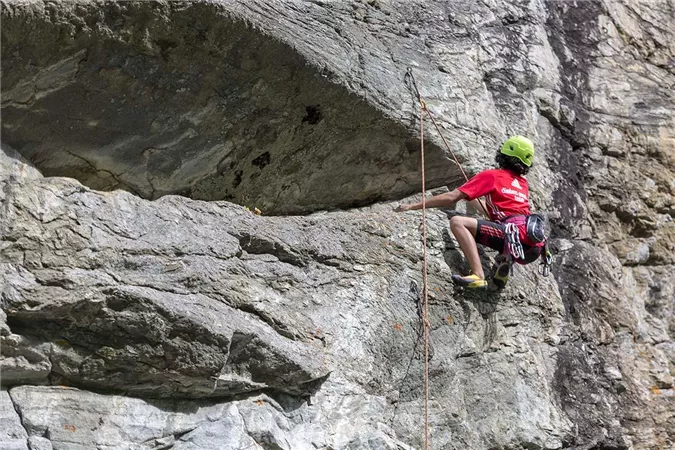 Ein Kletterer in einem roten Shirt erklimmt einen steilen Felsen. Er trägt einen Helm und eine Kletterausrüstung.