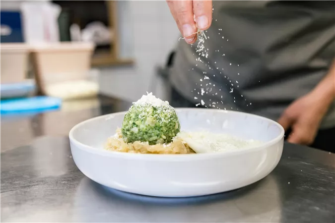 A plate with a green, ball-shaped dish is sprinkled with cheese. In the background, more kitchen utensils are visible.