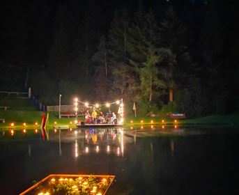 Eine stimmungsvolle Nachtaufnahme eines Sees mit beleuchtetem Floß. Im Hintergrund sind Bäume und eine sanfte Lichtquelle zu sehen.