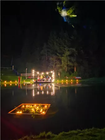 Eine stimmungsvolle Nachtaufnahme eines Sees mit beleuchtetem Floß. Im Hintergrund sind Bäume und eine sanfte Lichtquelle zu sehen.