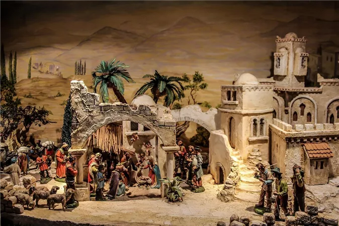 A detailed nativity scene with figures depicting the birth of Jesus. In the background, buildings and trees can be seen in a rural landscape.
