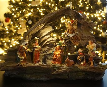 Eine weihnachtliche Krippe mit Figuren, die die Geburt Jesu darstellen. Im Hintergrund sind festlich beleuchtete Tannenbäume zu sehen.