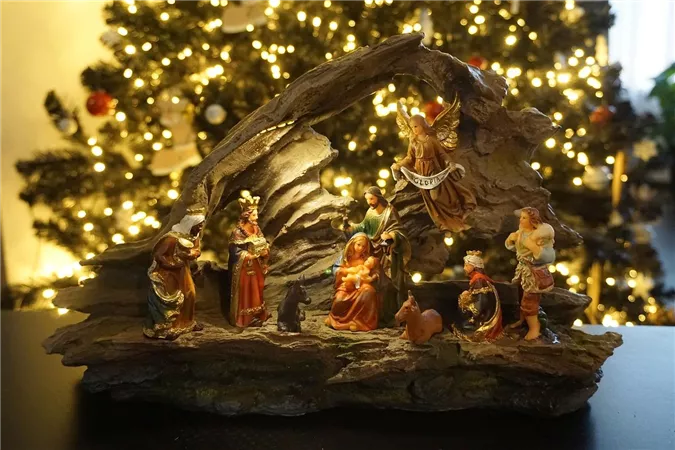 Eine weihnachtliche Krippe mit Figuren, die die Geburt Jesu darstellen. Im Hintergrund sind festlich beleuchtete Tannenbäume zu sehen.
