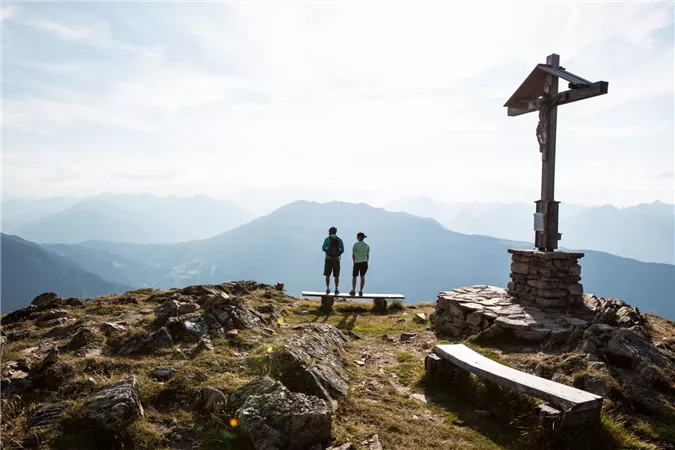 Zwei Menschen stehen auf einem Berggipfel neben einem Kreuz. Im Hintergrund erstreckt sich eine beeindruckende Berglandschaft mit einem klaren Himmel.
