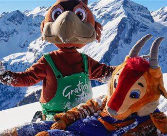 Zwei Maskottchen stehen im Schnee vor schneebedeckten Bergen. Eines der Maskottchen liegt auf dem Boden und das andere zeigt lächelnd seine Arme.