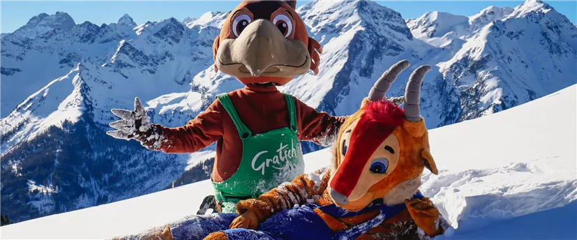 Zwei Maskottchen stehen im Schnee vor schneebedeckten Bergen. Eines der Maskottchen liegt auf dem Boden und das andere zeigt lächelnd seine Arme.
