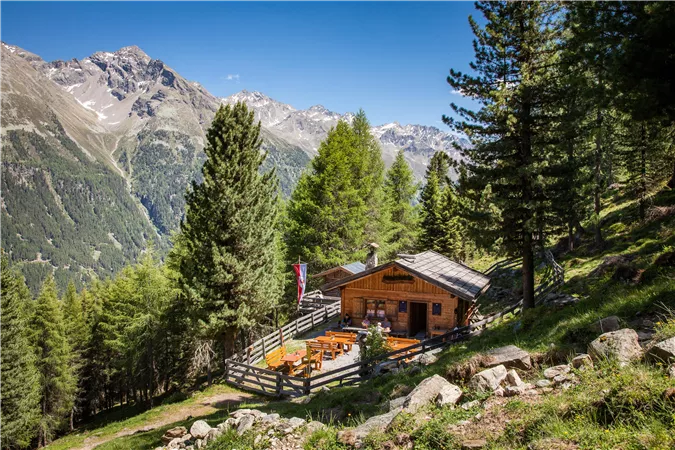 Eine gemütliche Alm umgeben von hohen Bäumen und Bergen. Die Terrasse lädt zum Entspannen in der Natur ein.