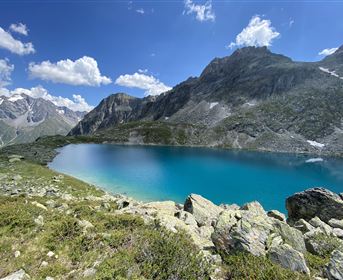 A clear, turquoise lake surrounded by tall mountains and green grass. The sky is blue with a few white clouds.