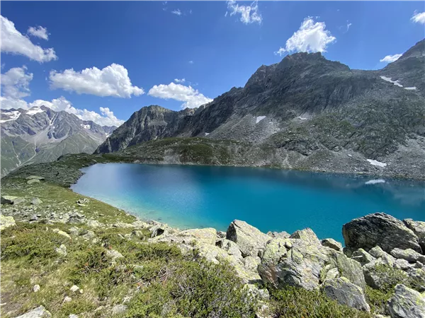Ein klarer, turquoise See umgeben von hohen Bergen und grünem Gras. Der Himmel ist blau mit einigen weißen Wolken.