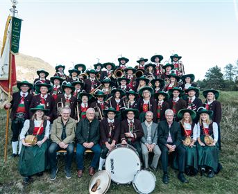 A large group of people in traditional outfits stands in front of a picturesque mountain landscape. Some people sit in the foreground while others stand in the second row.