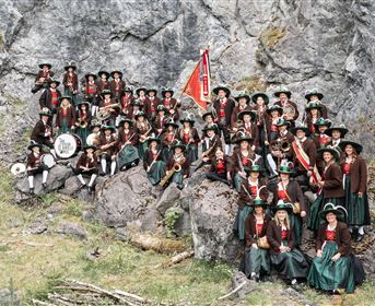 Eine große Gruppe von Menschen in traditionellen Trachten steht vor einer Felswand. Sie halten Musikinstrumente und eine Fahne und lächeln freundlich.