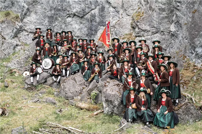 Eine große Gruppe von Menschen in traditionellen Trachten steht vor einer Felswand. Sie halten Musikinstrumente und eine Fahne und lächeln freundlich.