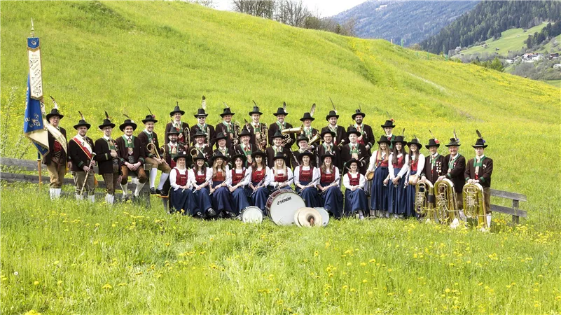 A large group of musicians stands in a green field. They are wearing traditional costumes and holding musical instruments.