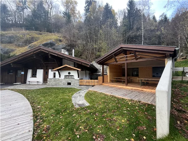 Zwei moderne Holzhütten in einer natürlichen Umgebung. Im Vordergrund liegt eine grüne Wiese mit vereinzelten herbstlichen Blättern.