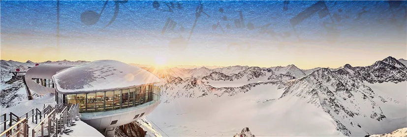 A modern mountain station with snow is situated in a beautiful Alpine landscape. In the background, majestic mountains rise under a clear sky.
