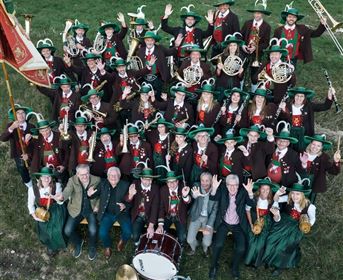 A large brass band is outdoors and smiling at the camera. Everyone is wearing green hats and festive clothing.