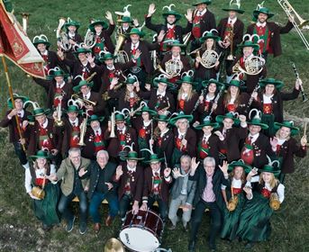 A large brass band with many members is standing outdoors. The musicians are wearing green hats and festive clothing and appear cheerful.