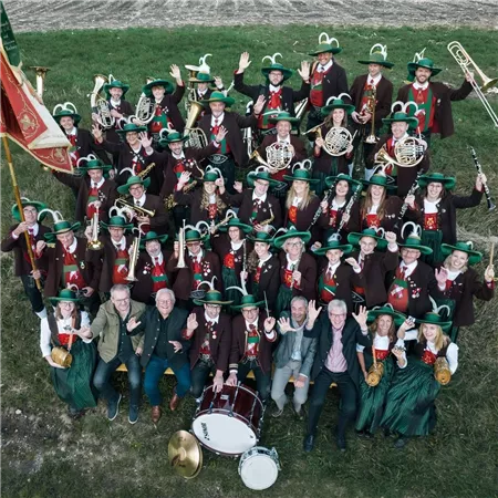 Eine große Blaskapelle mit vielen Mitgliedern steht im Freien. Die Musiker tragen grüne Hüte und festliche Kleidung und zeigen sich fröhlich.