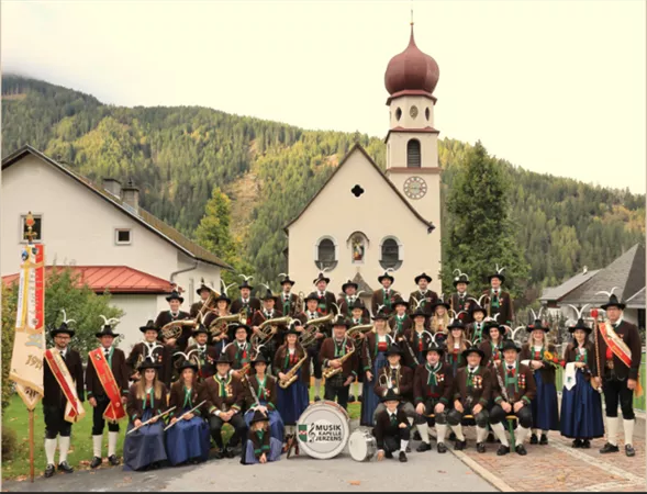 A large group of musicians in traditional costumes stands in front of a church and picturesque mountains. They are holding instruments and showing enthusiasm for their music.