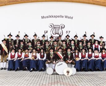 A music group in traditional attire stands in front of a wall with the inscription "Musikapelle Wald". All members are smiling and holding instruments in their hands.