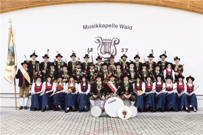 A music group in traditional attire stands in front of a wall with the inscription "Musikapelle Wald". All members are smiling and holding instruments in their hands.