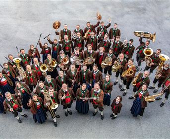 A large brass band stands together and holds their instruments in the air. All members wear traditional costumes and smile at the camera.