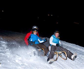 Zwei Personen fahren mit dem Schlitten die verschneite Piste hinunter. Es ist Nacht, und etwas Licht erhellt die Szene.