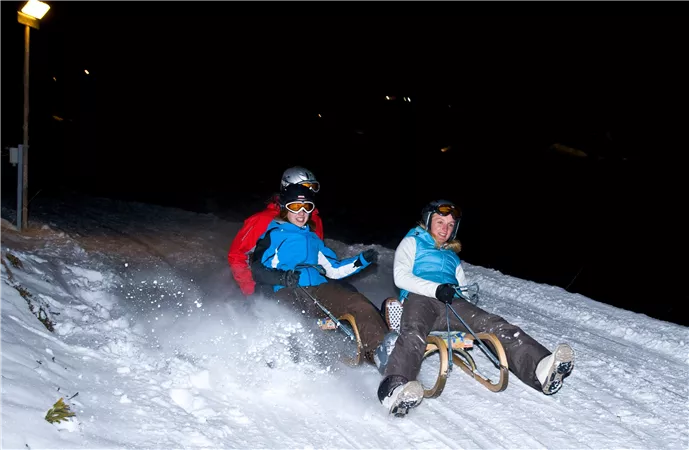 Zwei Personen fahren mit dem Schlitten die verschneite Piste hinunter. Es ist Nacht, und etwas Licht erhellt die Szene.