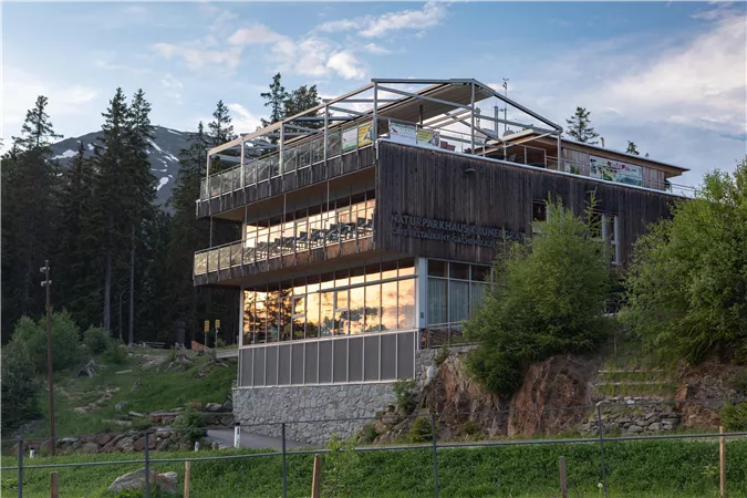 A modern building in the mountains with large windows. It is surrounded by trees and a natural landscape.