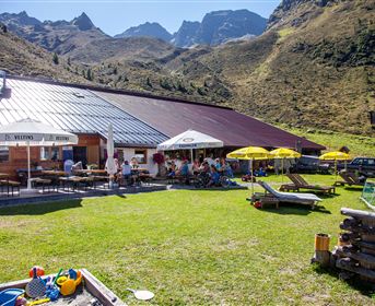 A cozy restaurant in the mountains with an inviting terrace. In the background, green hills and tall mountains can be seen.