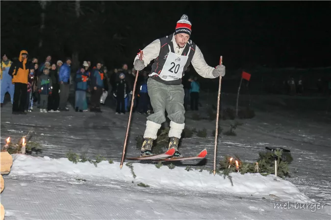 Ein Skifahrer springt über eine kleine Rampe im Schneeleuchten der Nacht. Zuschauer stehen im Hintergrund und beobachten das Ereignis.