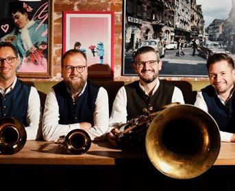 Four musicians are smiling behind a table, holding various wind instruments. In the background, colorful images and a city view can be seen.