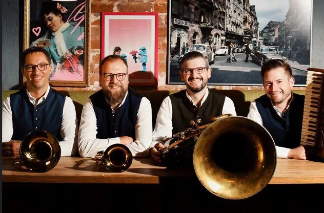 Four musicians are smiling behind a table, holding various wind instruments. In the background, colorful images and a city view can be seen.