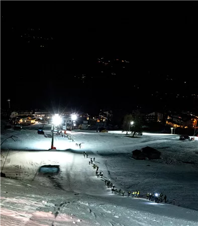 Eine schneebedeckte Piste bei Nacht mit beleuchteten Skiliften. Die Umgebung ist ruhig und es sind einige Menschen unterwegs.