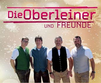 Eine Gruppe von vier Männern steht nebeneinander. Sie tragen traditionelle Kleidung und lächeln vor einem hellen, festlichen Hintergrund.