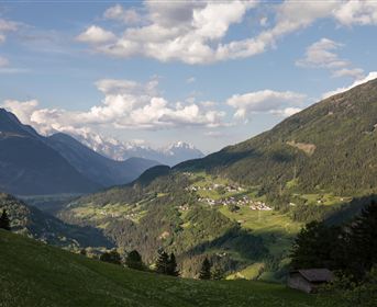 A picturesque mountain landscape with green meadows and gentle hills. In the background, there are high mountains and a blue sky with some clouds.