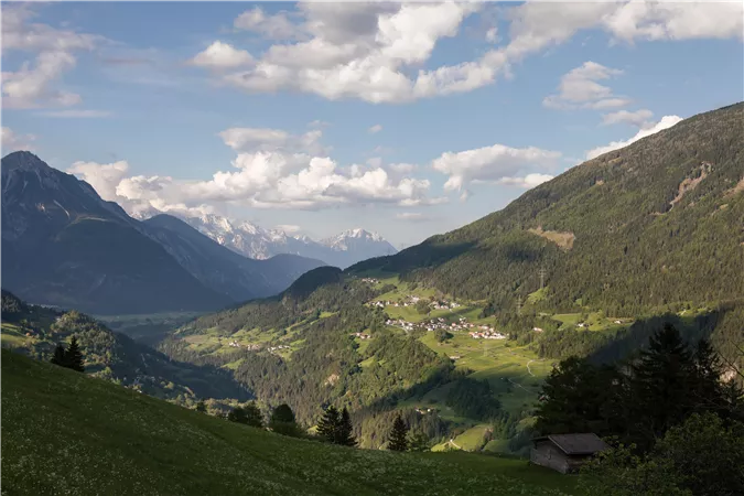 A picturesque mountain landscape with green meadows and gentle hills. In the background, there are high mountains and a blue sky with some clouds.