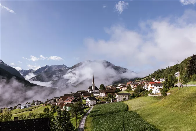 A picturesque mountain landscape with clouds and a small town. In the foreground are lush meadows and houses.