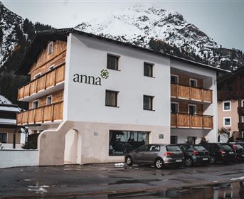 Ein modernes Gebäude mit Holzverkleidung und dem Schriftzug „anna“. Im Hintergrund sind schneebedeckte Berge zu sehen.