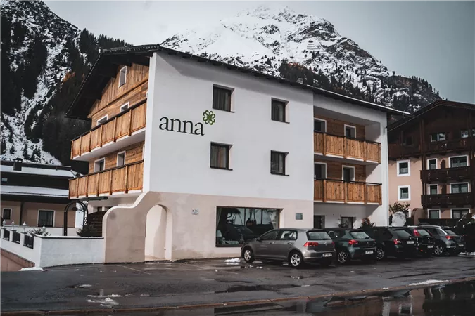 Ein modernes Gebäude mit Holzverkleidung und dem Schriftzug „anna“. Im Hintergrund sind schneebedeckte Berge zu sehen.