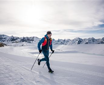 Ein Mann läuft auf einer schneebedeckten Berglandschaft. Im Hintergrund sind hohe Berge und ein klarer Himmel zu sehen.