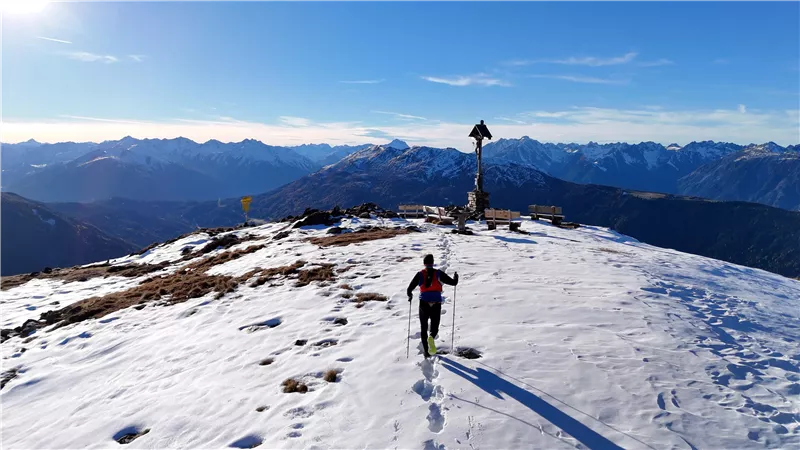 Eine schneebedeckte Bergspitze mit klaren blauen Himmel. Eine Person wandert mit Skiern in die Richtung eines Gipfelkreuzes.