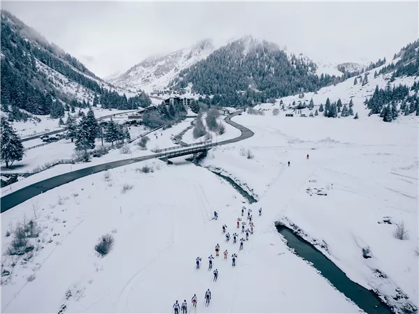 Eine verschneite Landschaft mit Bergen im Hintergrund. Gruppen von Menschen wandern entlang eines verschneiten Weges neben einem Fluss.