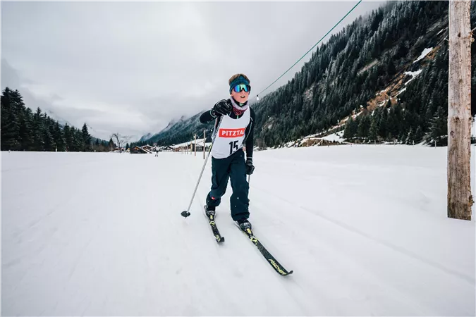 Ein Skifahrer fährt auf einer verschneiten Piste. Im Hintergrund sind verschneite Berge und bewaldete Hügel zu sehen.