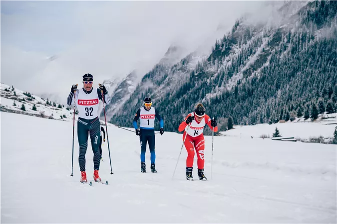 Drei Langläufer in einem schneebedeckten Landschaftsbereich in den Bergen. Der Himmel ist bewölkt und die Umgebung ist winterlich.